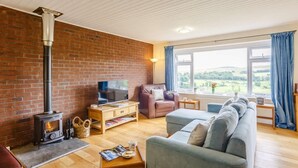 A cozy living room with a brick wall, a fireplace, a sofa, a TV, and a window with a view. - 3 Bed in Morpeth (oc-63025) (Rothbury)