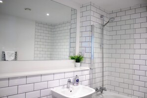 Un bagno con pareti piastrellate di bianco, un lavandino bianco, cabina doccia in vetro e una piccola pianta in vaso sul mobile.