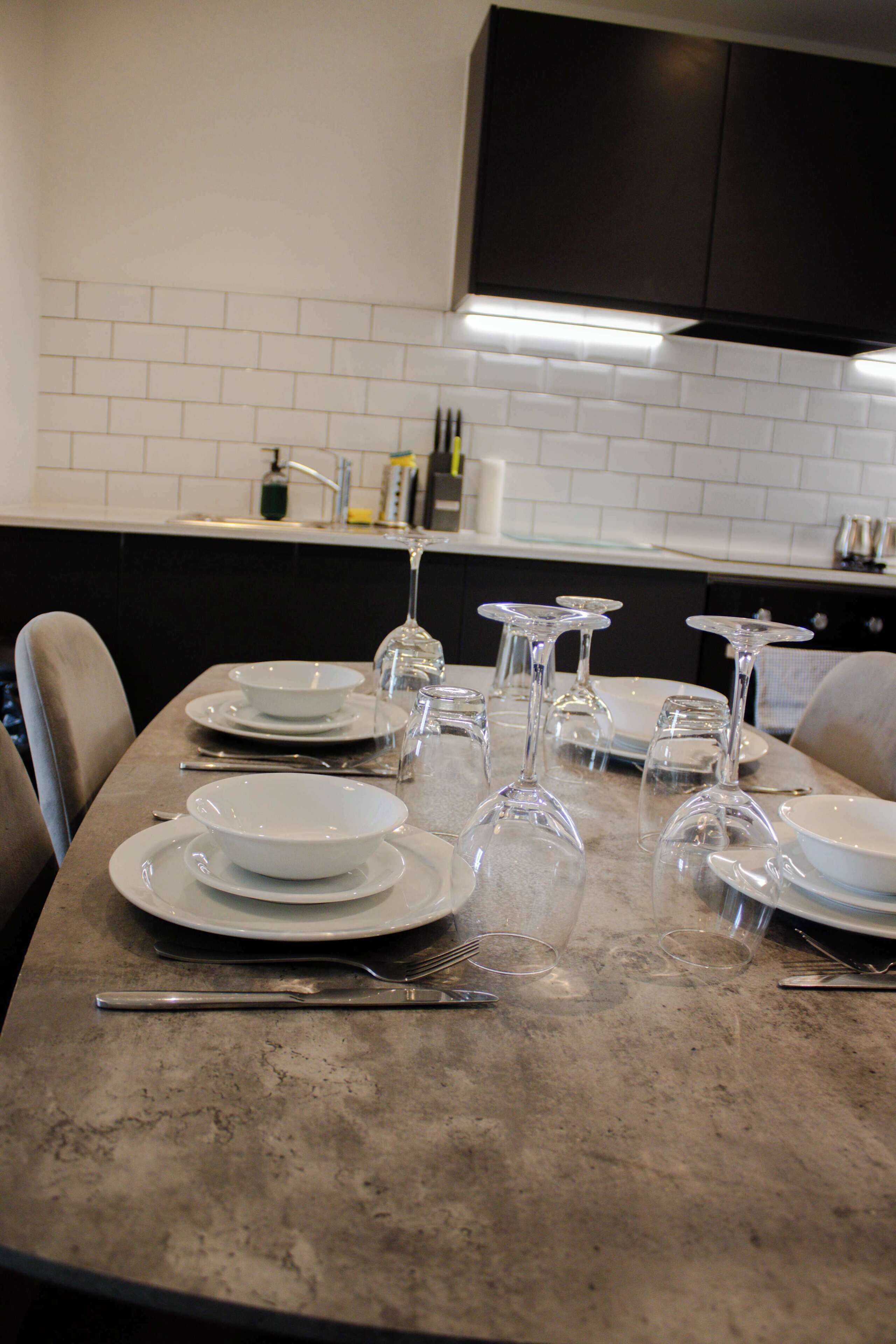 Een eettafel gedekt met borden, glazen en bestek in een moderne keuken met witte tegels en donkere kastjes.