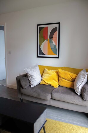 A modern living room with a grey sofa, yellow and white cushions, a black coffee table, and a framed abstract painting on the wall. - Effortless Stays at 2BR Escape OldTrafford Stadium (Stretford)