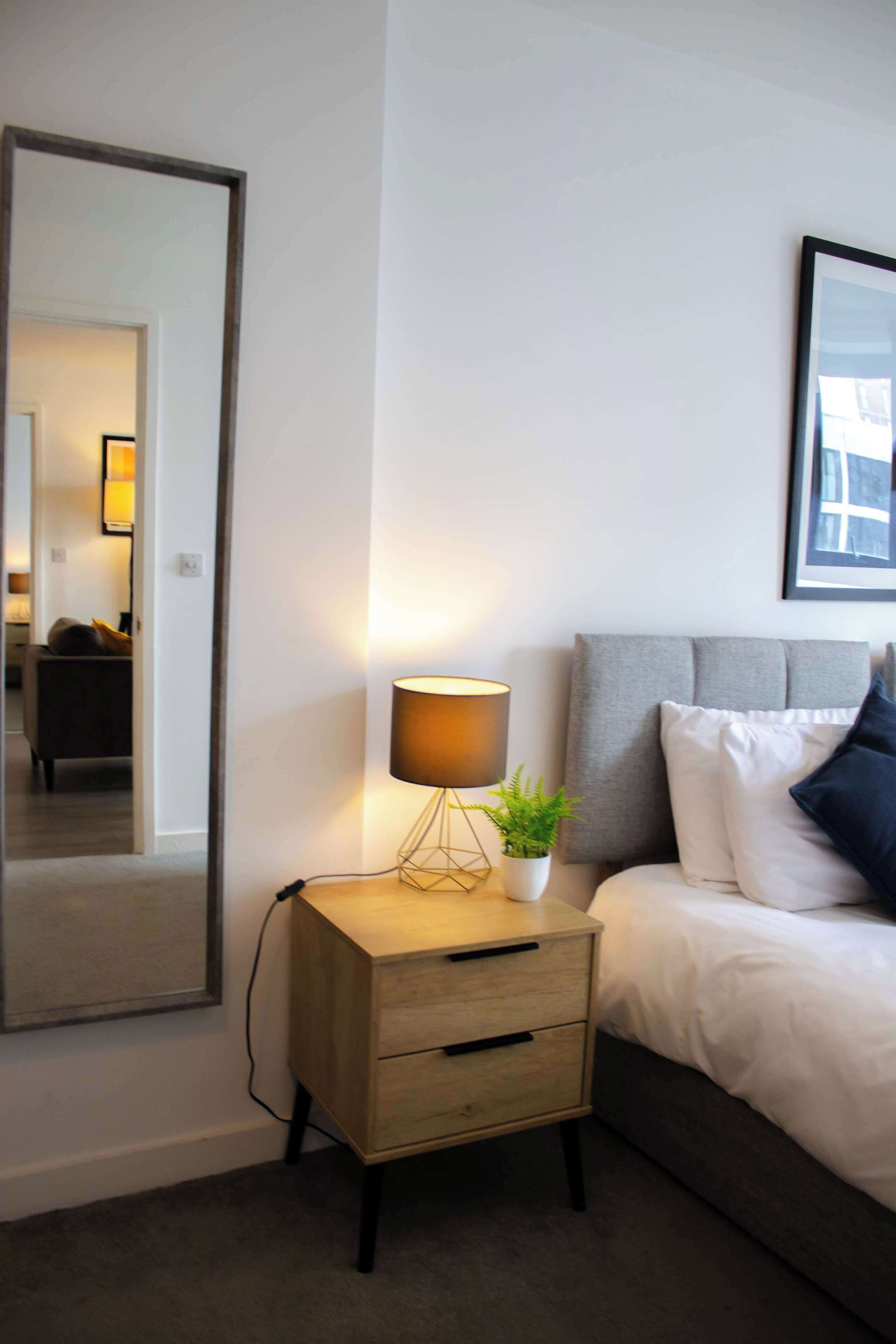 Quarto com uma cama, abajur, uma pequena mesa de cabeceira de madeira, um espelho e um quadro na parede.