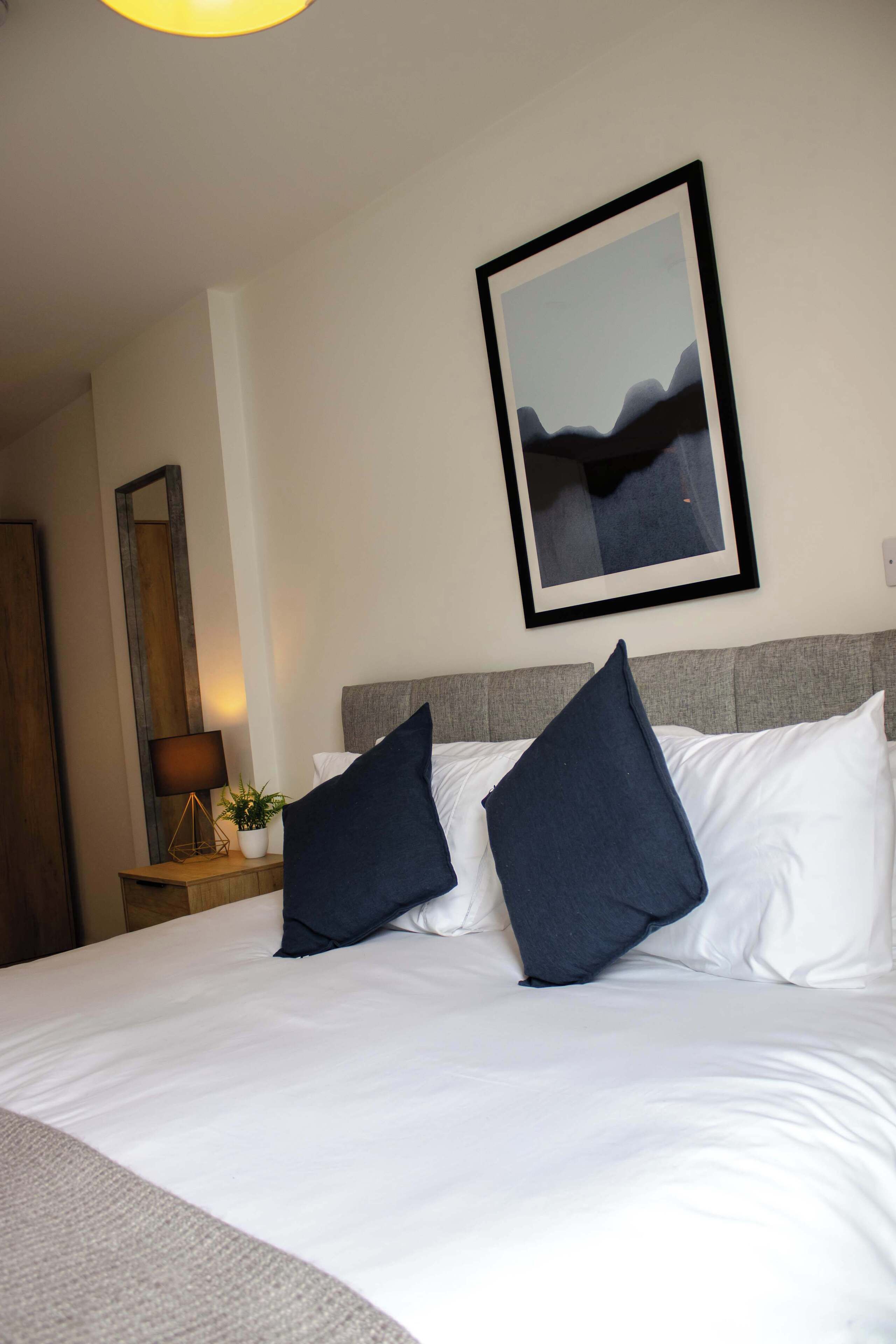 A hotel room with a neatly made bed, a bedside lamp, a framed picture on the wall, and a wooden nightstand with a plant.