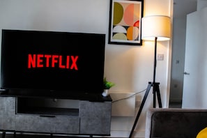 A modern living room with a large flat-screen TV displaying 'NETFLIX,' a floor lamp, and a framed abstract art piece on the wall. - Effortless Stays at 2BR Escape OldTrafford Stadium (Stretford)