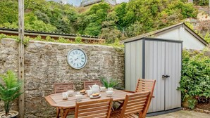 Un patio con tavolo e sedie, una sveglia su un muro in pietra e un piccolo deposito.
