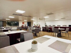 A conference room with multiple tables, chairs, a large screen, and a kitchenette area.