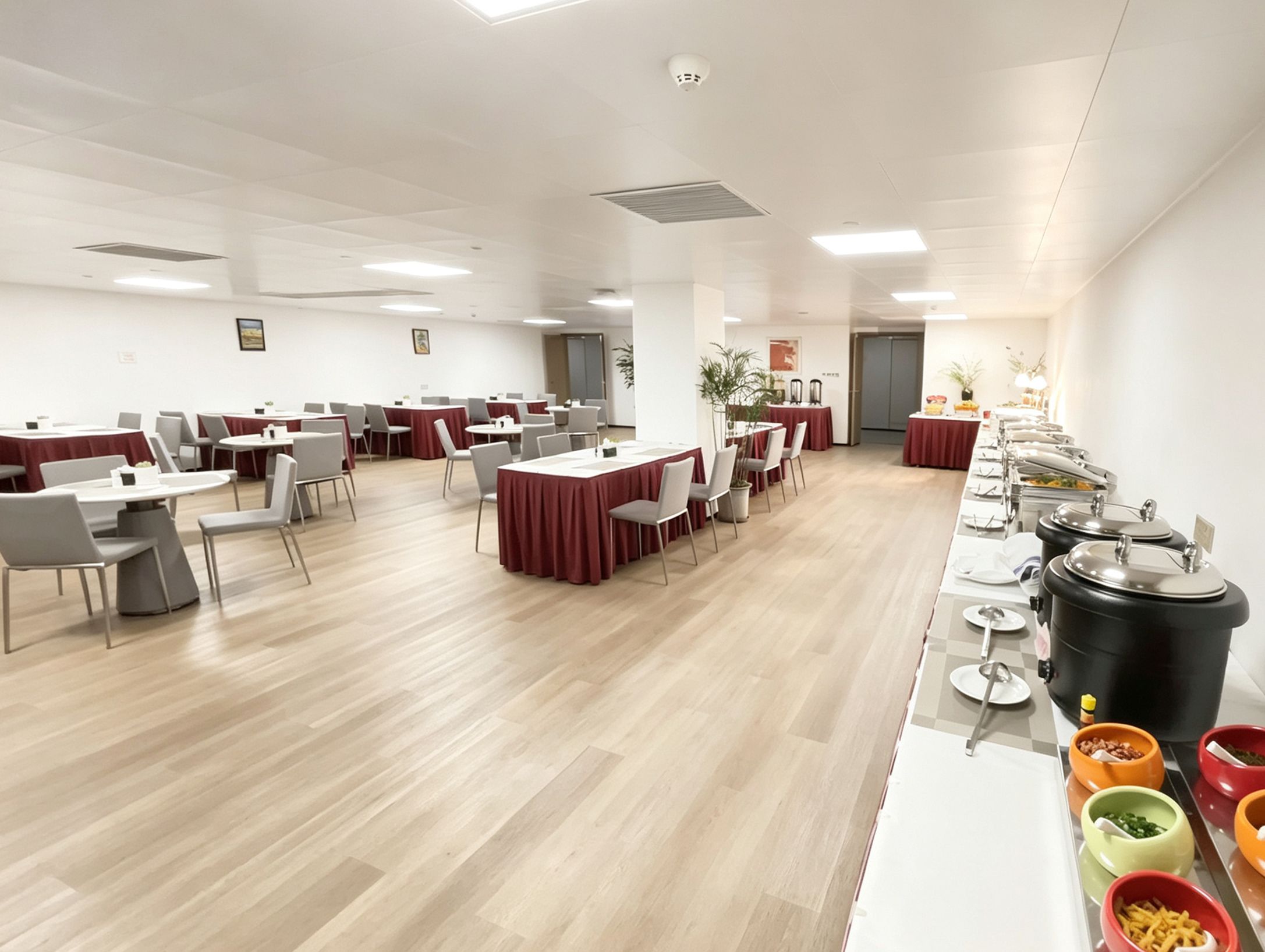 A spacious conference room with multiple tables and chairs, equipped with a buffet line on one side.