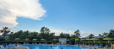 A swimming pool with lounge chairs and umbrellas, surrounded by greenery and buildings.