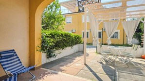 Een overdekt buitengebied met terras, een lounge stoel met blauwe strepen, een kleine tafel en een pergola met witte gordijnen.