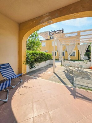 Ein überdachter Außenbereich mit Terrasse, einem blau gestreiften Liegestuhl, einem kleinen Tisch und einer Pergola mit weißen Vorhängen.