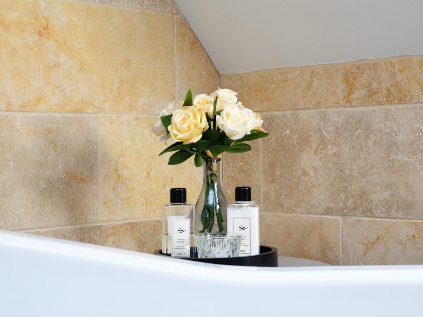 A bathroom with a marble wall, a white bathtub, a vase with white flowers, and two bottles of bath products.