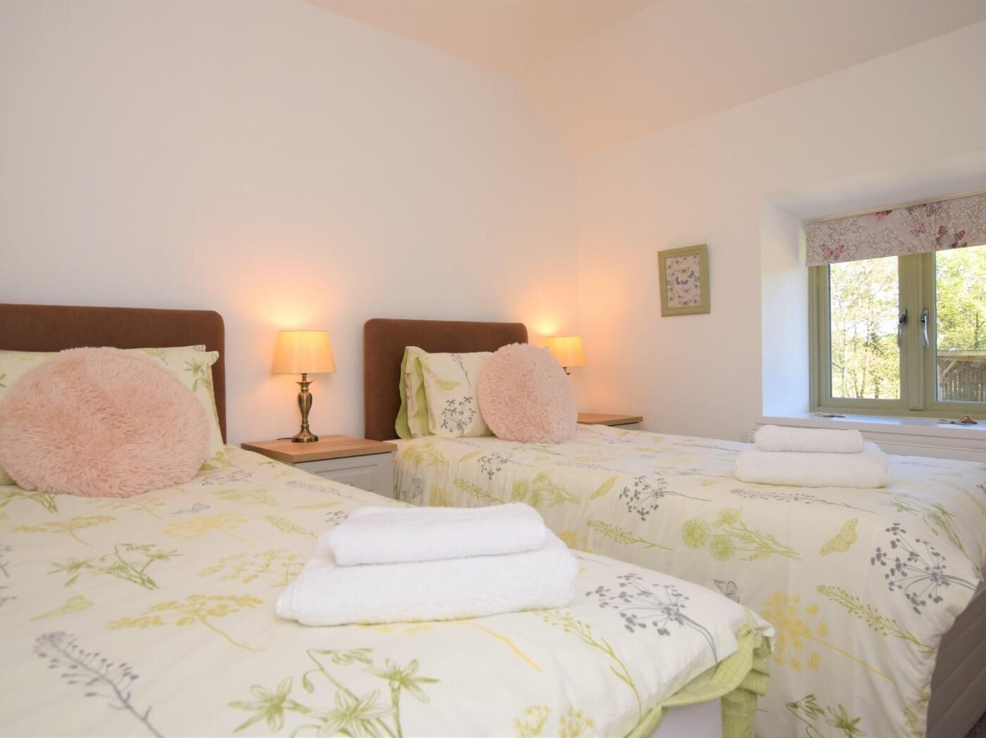 A room with two beds, each with a pillow and a lamp. There is a window with a view of trees.