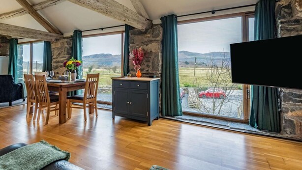 Un espace repas comprenant une table, des chaises et offrant une vue sur la campagne grâce à de grandes fenêtres.
