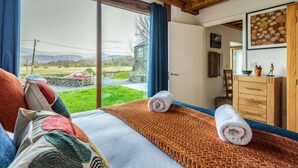 Une chambre avec une grande fenêtre, des poutres apparentes, un lit avec des serviettes roulées, une commode et une vue sur un paysage rural.
