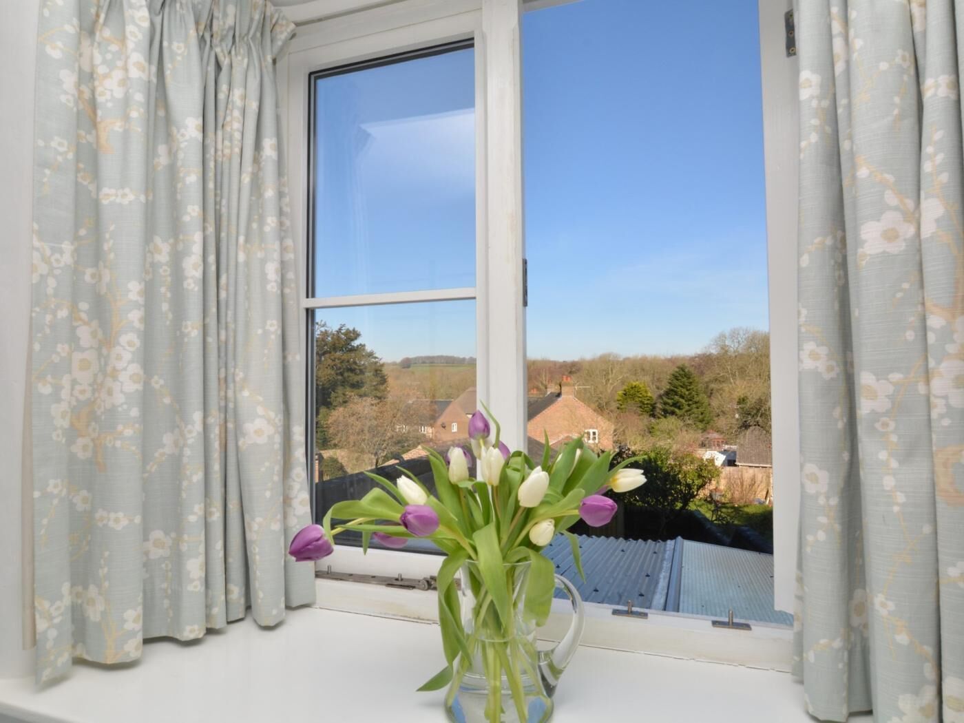 Una ventana con vistas a un paisaje rural, un florero con flores y cortinas.