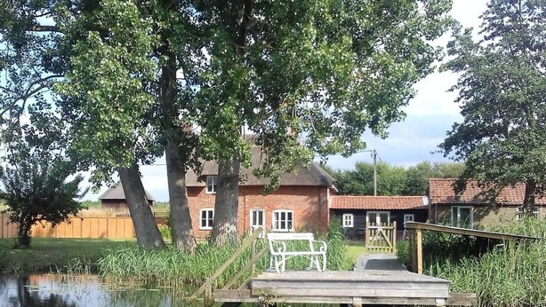 A riverside property with a wooden dock, brick buildings, and a grassy area. - 6 Bed in Norwich (oc-72652) (Hoveton)