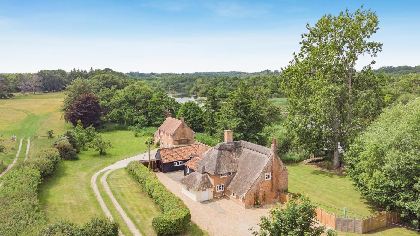 A rural property with a thatched-roof house, a fenced yard, and a river in the background.