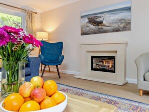 A living room with a fireplace, a blue armchair, a vase with flowers, and a bowl of fruit. - 5 Bed in Woodbridge (oc-72724) (Woodbridge)