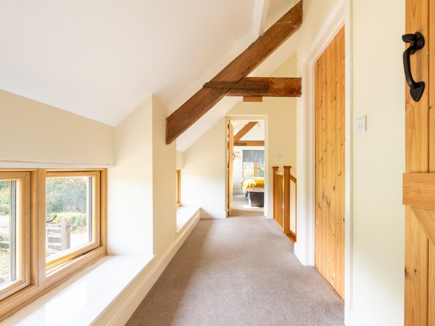 A hallway with wooden beams, a carpeted floor, and a view into a bedroom with a bed and a window.