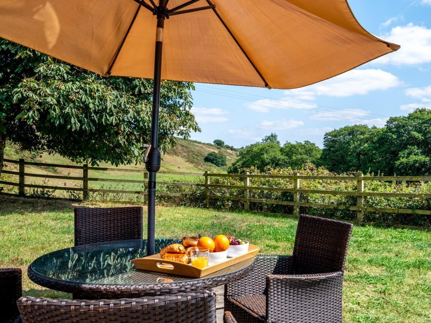 A round glass table with wicker chairs, an umbrella, and a tray of food.