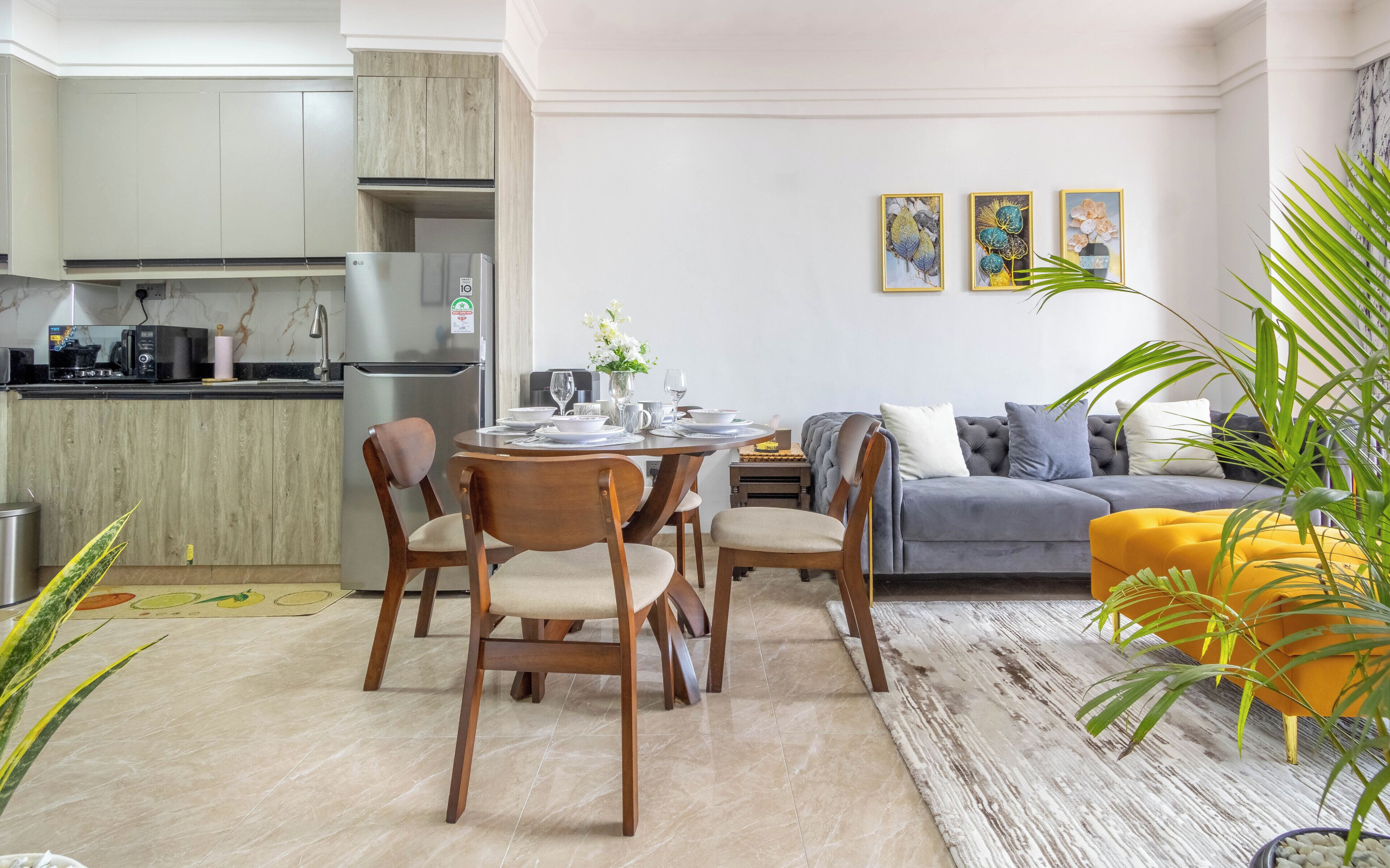 Una cocina moderna con un refrigerador LG empotrado, un comedor con sillas de madera, un sofá gris y un reposapiés amarillo.