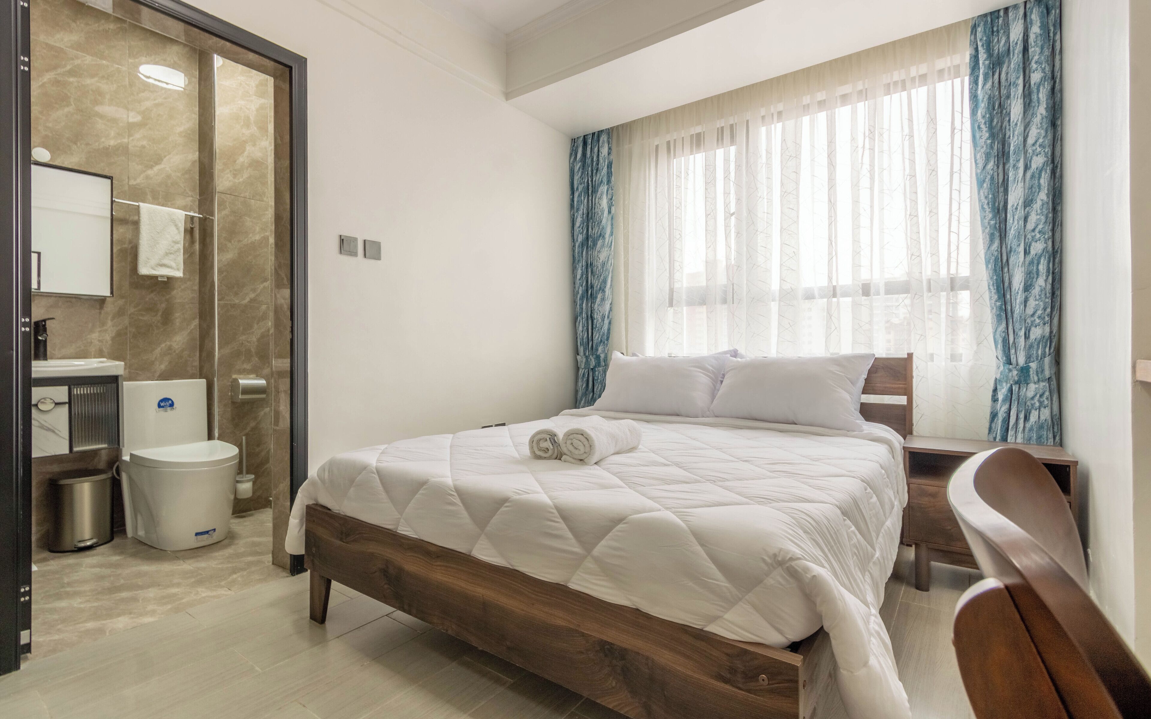 Habitación de hotel con cama, mesita de noche de madera, silla, baño con ducha y ventana con cortinas.