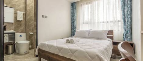 Habitación de hotel con cama, mesita de noche de madera, silla, baño con ducha y ventana con cortinas.