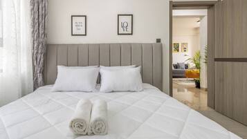 Una cama bien tendida con toallas blancas enrolladas, un cabecero tapizado de color gris y dos citas inspiradoras enmarcadas en la pared.