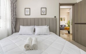 Ein ordentlich bezogenes Bett mit aufgerollten weißen Handtüchern, einem grau gepolsterten Kopfteil und zwei gerahmten Motivationssprüchen an der Wand.