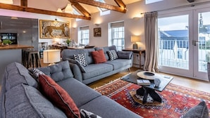Ein großzügiger Wohnbereich mit grauem Sofa, Couchtisch und einem Teppich mit rotem Muster. Es gibt eine Essecke mit Tisch und Stühlen sowie eine Küche mit modernen Geräten. Der Raum zeichnet sich durch freiliegende Holzbalken und große Fenster aus.