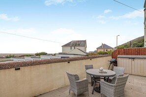 Eine Terrassengruppe im Freien mit rundem Tisch, Korbstühlen, einem Grill und Blick auf Häuser im Hintergrund.