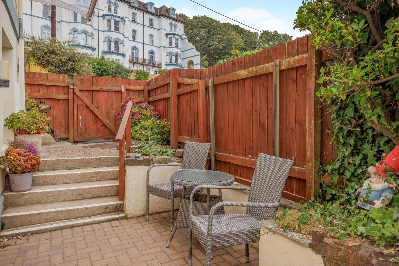 Une terrasse dotée de meubles en osier, d’une clôture en bois et d’un escalier menant à un bâtiment.