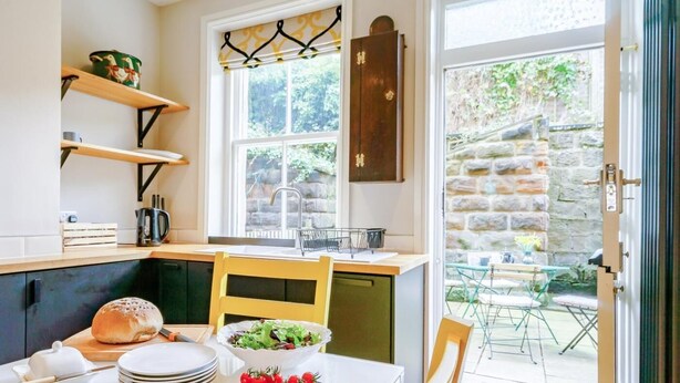 A kitchen with a window, a sink, a wooden table with plates, a bowl of salad, and a chair. - 4 Bed in Harrogate (oc-73424) (Harrogate)
