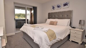 Ein Schlafzimmer mit einem Bett, einem Nachttisch, einer Lampe, einer Schiebeglastür zu einem Balkon und drei floralen Bildern, die an der Wand hängen.