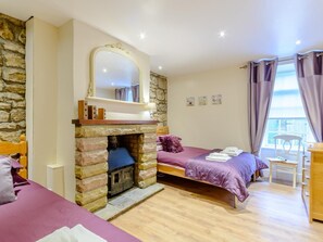 Ein Schlafzimmer mit Kamin aus Stein, einem Bett mit violettem Überwurf, einem Holzschrank und einem Fenster mit Vorhängen.