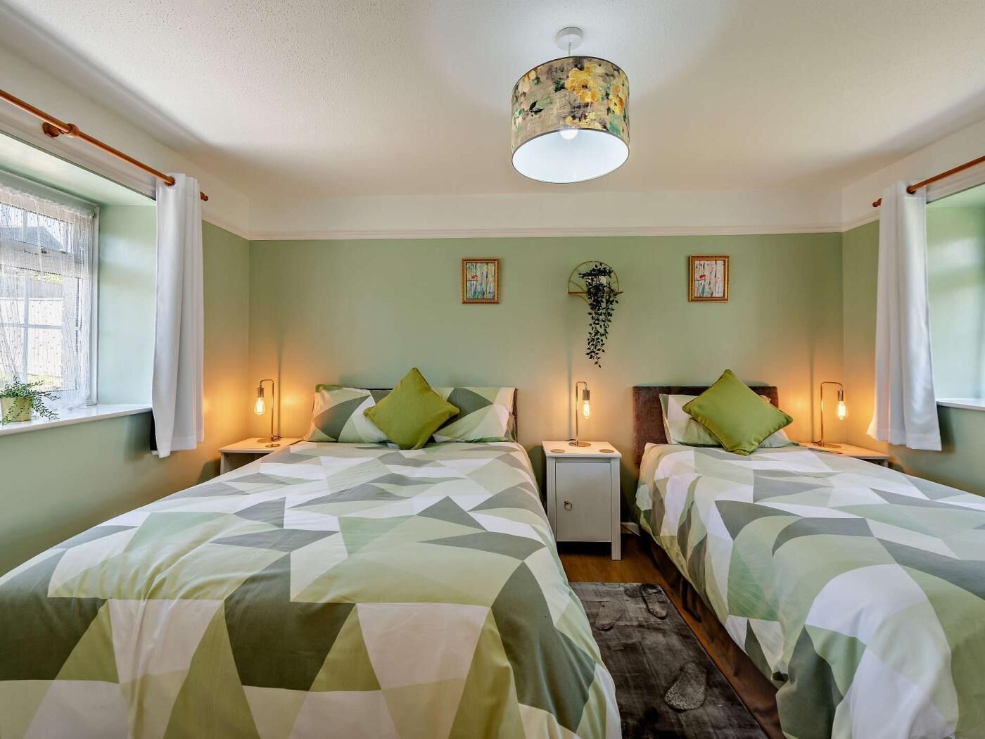 Une chambre avec deux lits, un luminaire suspendu, des tables de chevet avec des lampes et une fenêtre avec des rideaux.