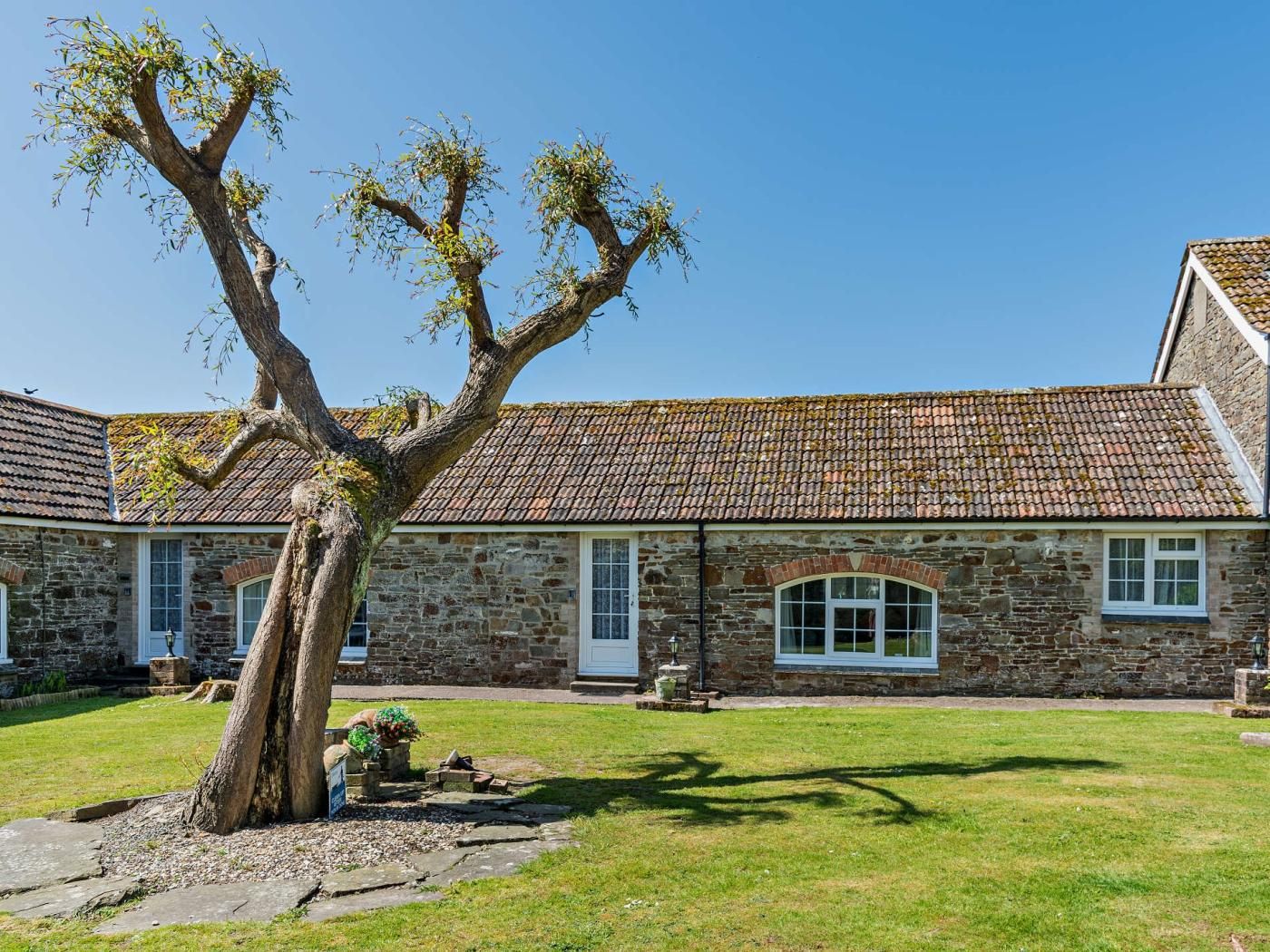 A stone building with a tiled roof, a large tree in the foreground, and a clear blue sky.