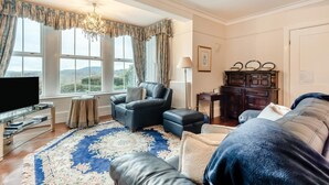 Ein Wohnzimmer mit einem großen Fenster, einem Fernseher, einem Ledersofa, einem Couchtisch und einem gemusterten Teppich.