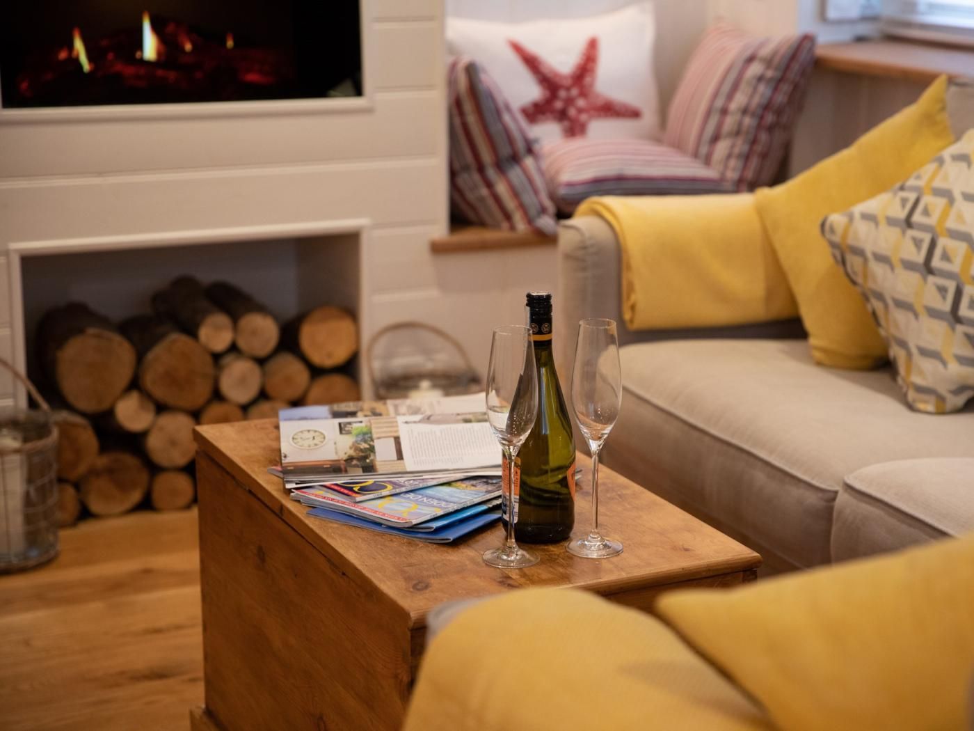 A cozy living room with a wooden coffee table holding a bottle and glasses, a fireplace with logs, and a sofa with patterned cushions.