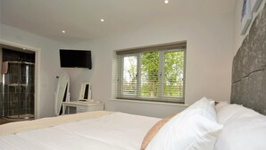 Quarto com cama, televisão fixada na parede, janela com persianas e área de chuveiro.