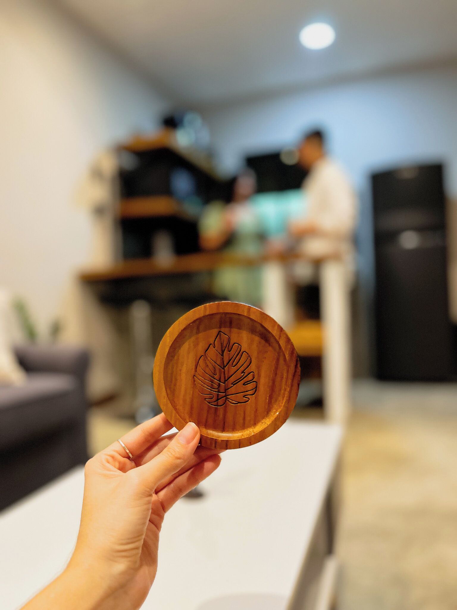 Una mano sostiene un posavasos de madera con diseño de hoja, una mesa blanca y un fondo desenfocado con una persona y un refrigerador.