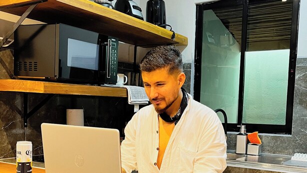 A person working on a laptop at a wooden desk in a kitchen with a microwave, coffee maker, and window. - Tropical Apartments (Puerto Viejo de Talamanca)