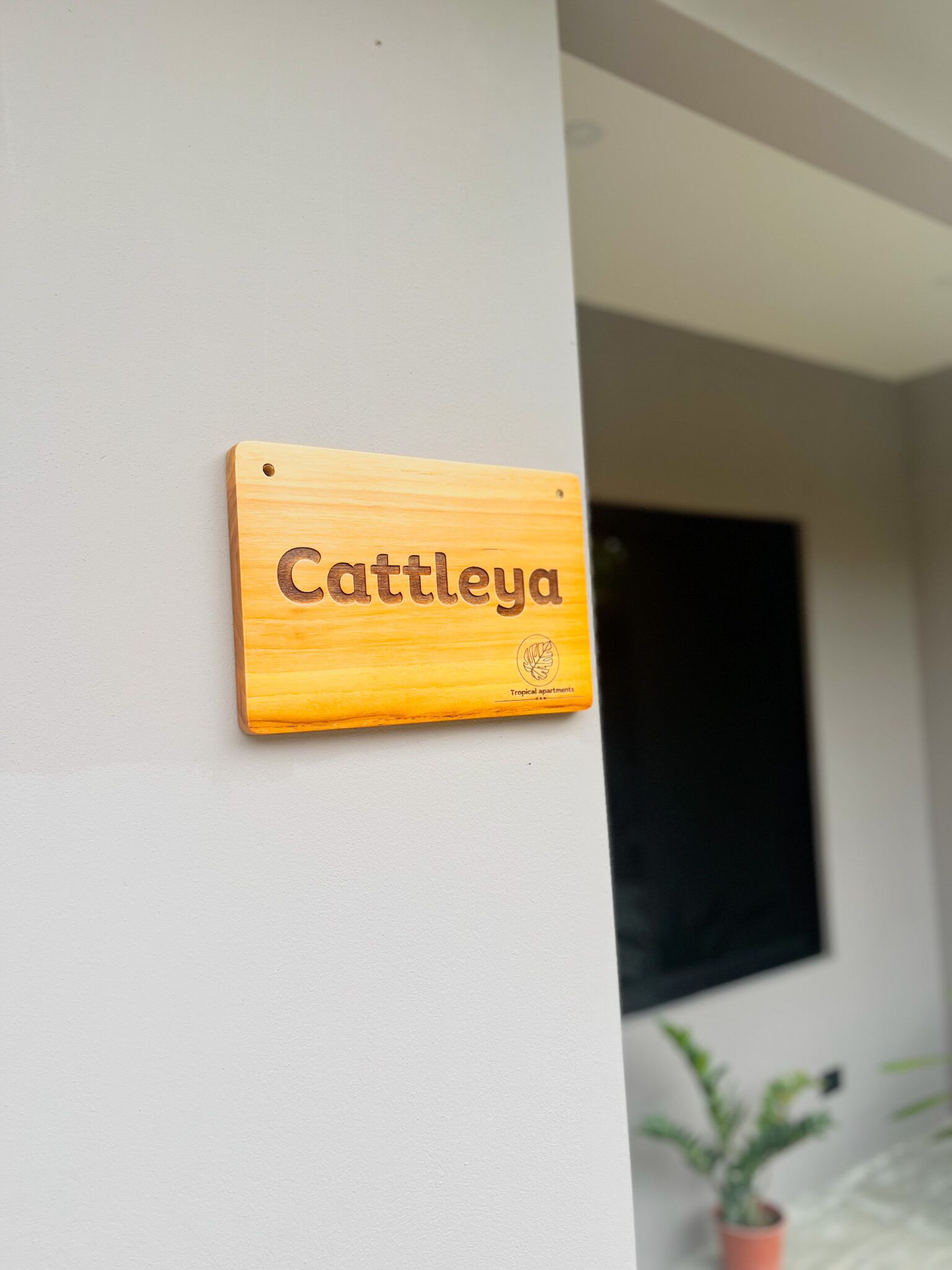 Une enseigne en bois portant le nom « Cattleya » et le logo de « Tropical apartments », fixée à un mur.