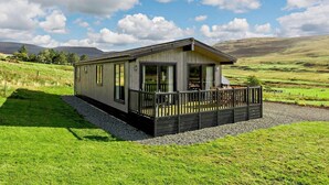 Une cabane en bois à un seul étage, dotée d’un balcon, entourée d’un paysage verdoyant et avec des collines en arrière-plan.