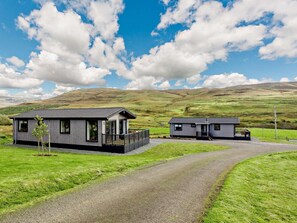 A modern two-story cabin with a balcony, situated in a rural landscape with rolling hills in the background.