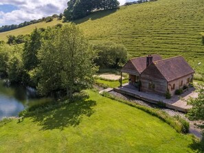 A rustic wooden house with a sloped roof, situated on a grassy hill with a river in the foreground. - 2 Bed in Dorchester (oc-75243) (Sydling Saint Nicholas)