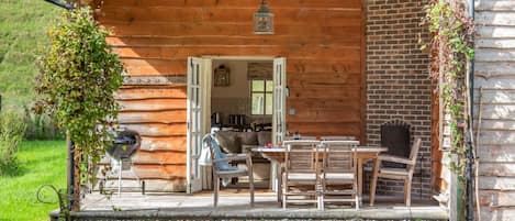 A wooden cabin with a patio area featuring a table and chairs, a grill, and a hanging lantern.