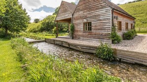 A rustic wooden house with a covered porch, a small window, and a gravel path leading to a pond. - 2 Bed in Dorchester (oc-75243) (Sydling Saint Nicholas)