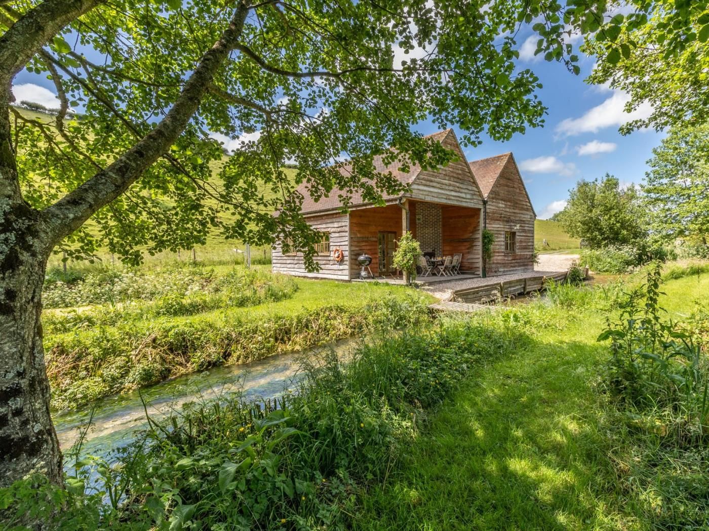 Un chalet rustique en bois, doté d’un porche, entouré d’une végétation luxuriante et bordé d’un petit ruisseau.