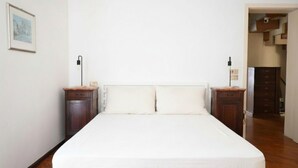 Une chambre à coucher avec un grand lit, des tables de chevet en bois, une lampe fixée au mur et une porte donnant sur un couloir.