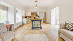 Een ruime woonkamer met een keuken op de achtergrond, een groot raam en een visgraatvloer.